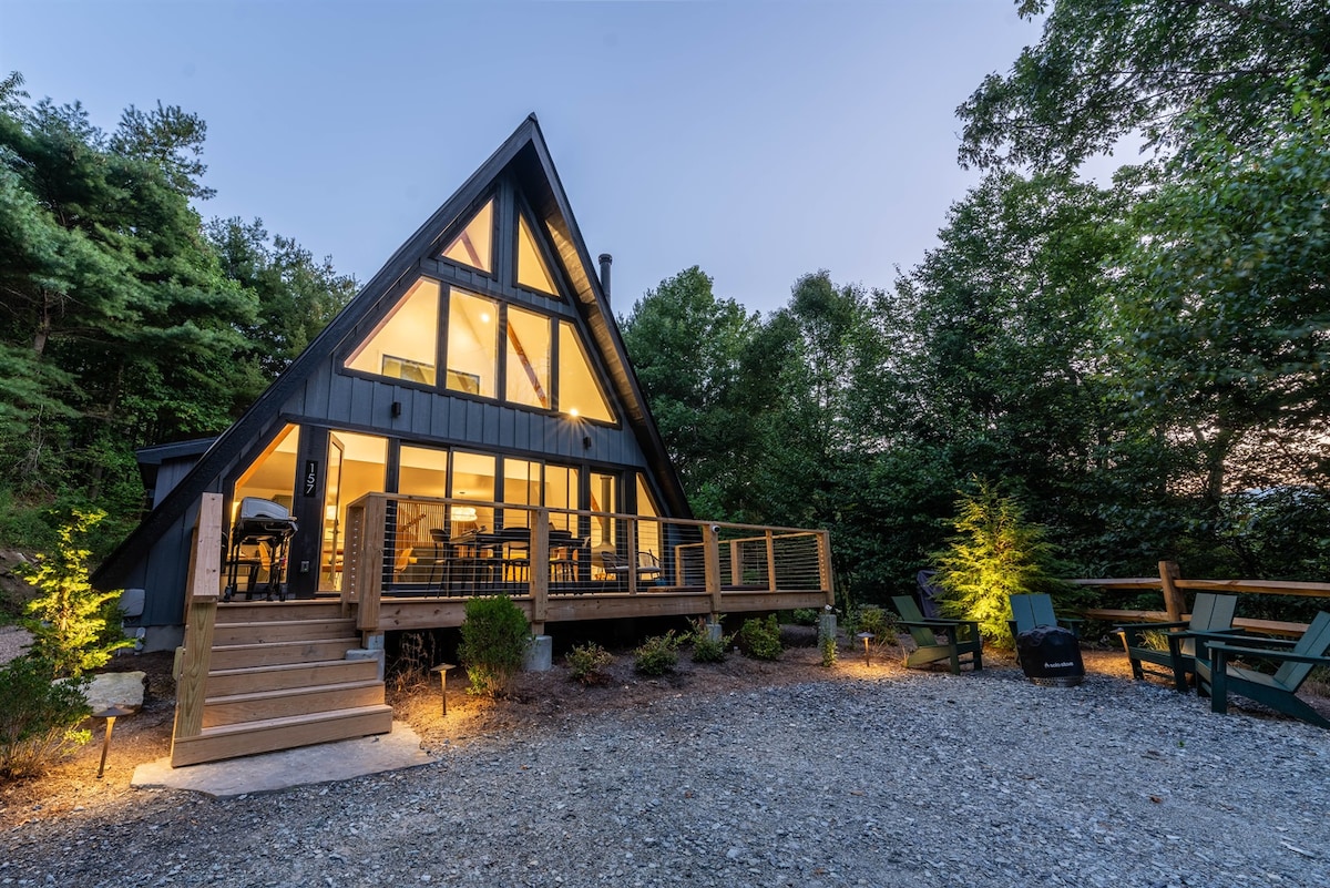 A modern A-Frame cabin is situated amidst lush greenery, illuminated by soft outdoor lighting. The structure features a deck with outdoor seating, and large floor-to-ceiling windows offer views of the interior space, creating a welcoming ambiance.