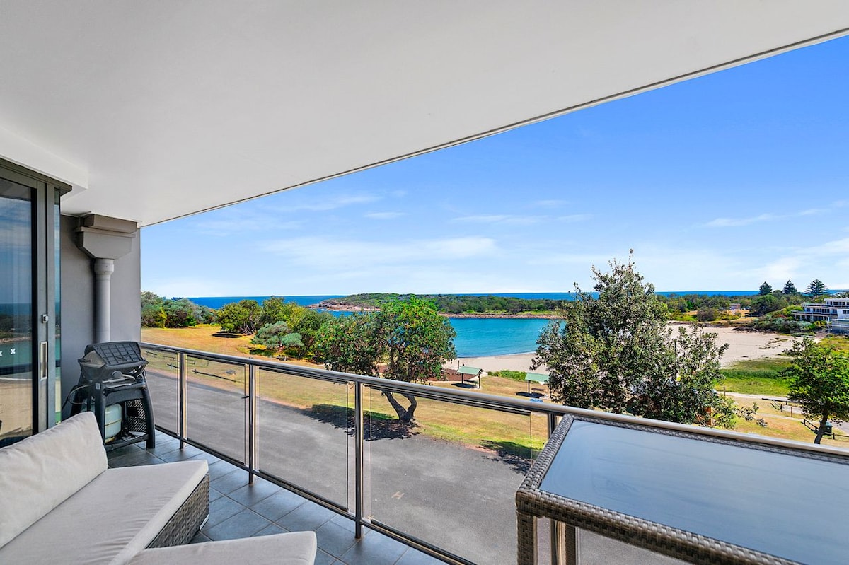 A spacious balcony showcases panoramic ocean views, with comfortable outdoor seating included. The calming blue water of Boat Harbour contrasts with the greenery of the nearby landscape, creating a relaxing coastal scene. A clear sky above enhances the light-filled open space.