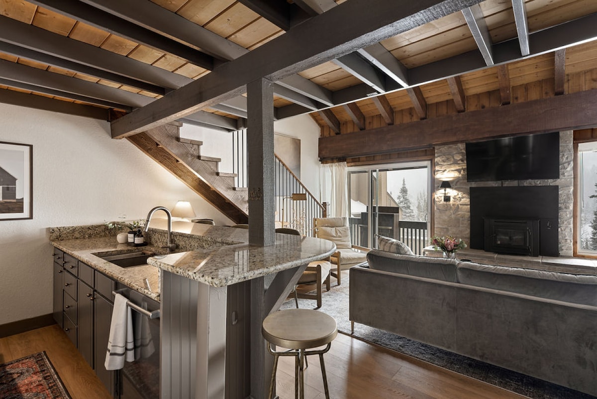 An open living space features a modern kitchen with granite countertops and a curved breakfast bar. The cozy seating area includes a soft couch, a large television mounted on a stone wall, and access to a deck with views of the surrounding nature.