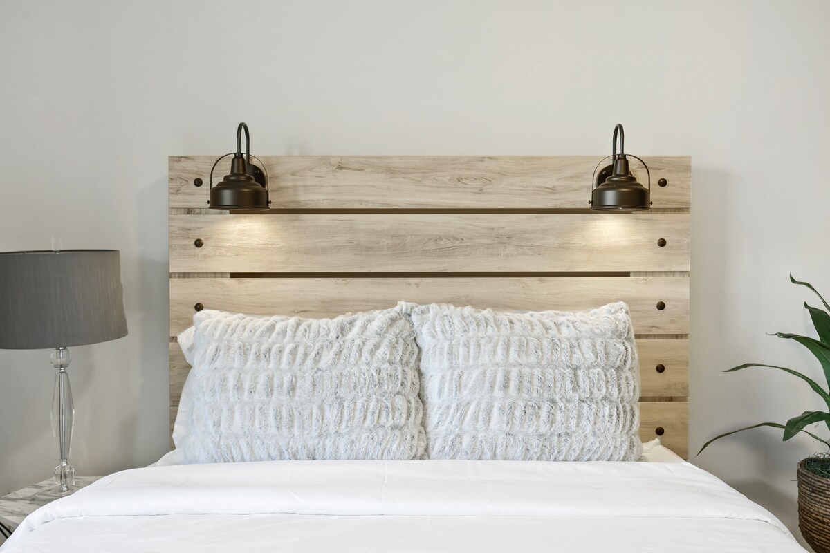 A well-appointed bedroom exhibits a wooden headboard with built-in lamps for illumination. Two plush pillows are placed against the headboard, complemented by a neatly arranged white bedspread. A bedside lamp and a potted plant add subtle touches of comfort.