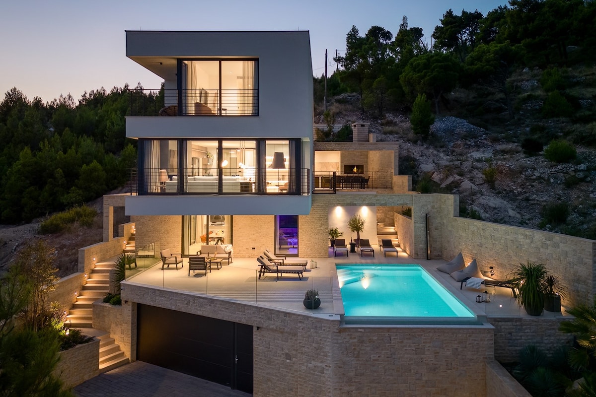 The villa is illuminated at dusk, showcasing a spacious terrace that includes a heated infinity pool. Lounge chairs are arranged around the pool, while outdoor seating areas are visible. Large glass doors connect the indoor spaces to the terrace, emphasizing the modern architecture.