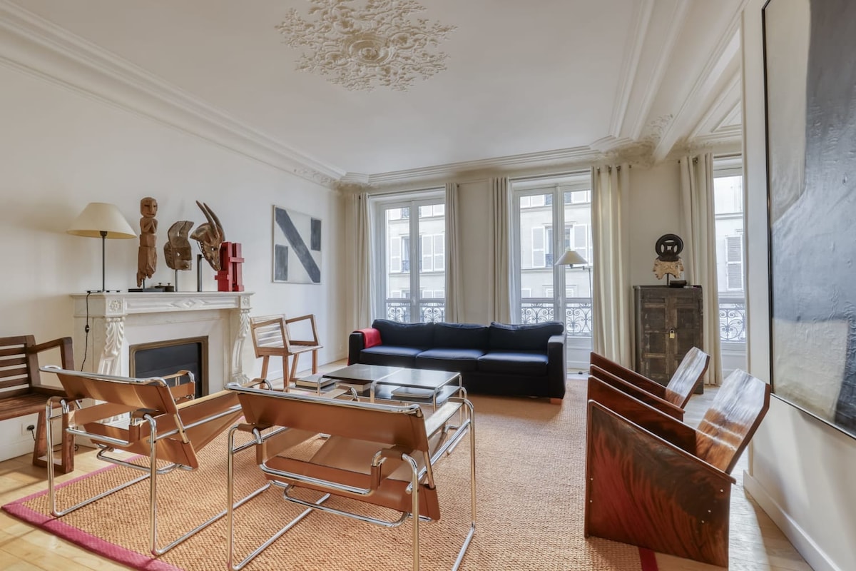 A spacious living room features a dark sofa and a central coffee table, surrounded by stylish wooden chairs. Natural light floods in through large windows, illuminating the warm-toned wooden floor and decorative artworks on the walls.