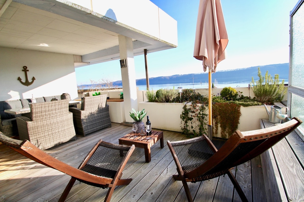 A private terrace features comfortable loungers and a stylish table, with views of the lake in the background. Soft sunlight illuminates the space, while greenery and decoration elements add to the relaxed ambiance.