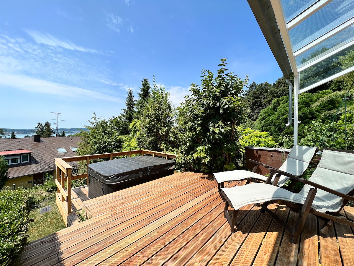 A spacious outdoor terrace features a wooden deck surrounded by greenery. A hot tub is positioned for scenic views, while two lounge chairs invite relaxation. The sky is clear, enhancing the serene atmosphere.