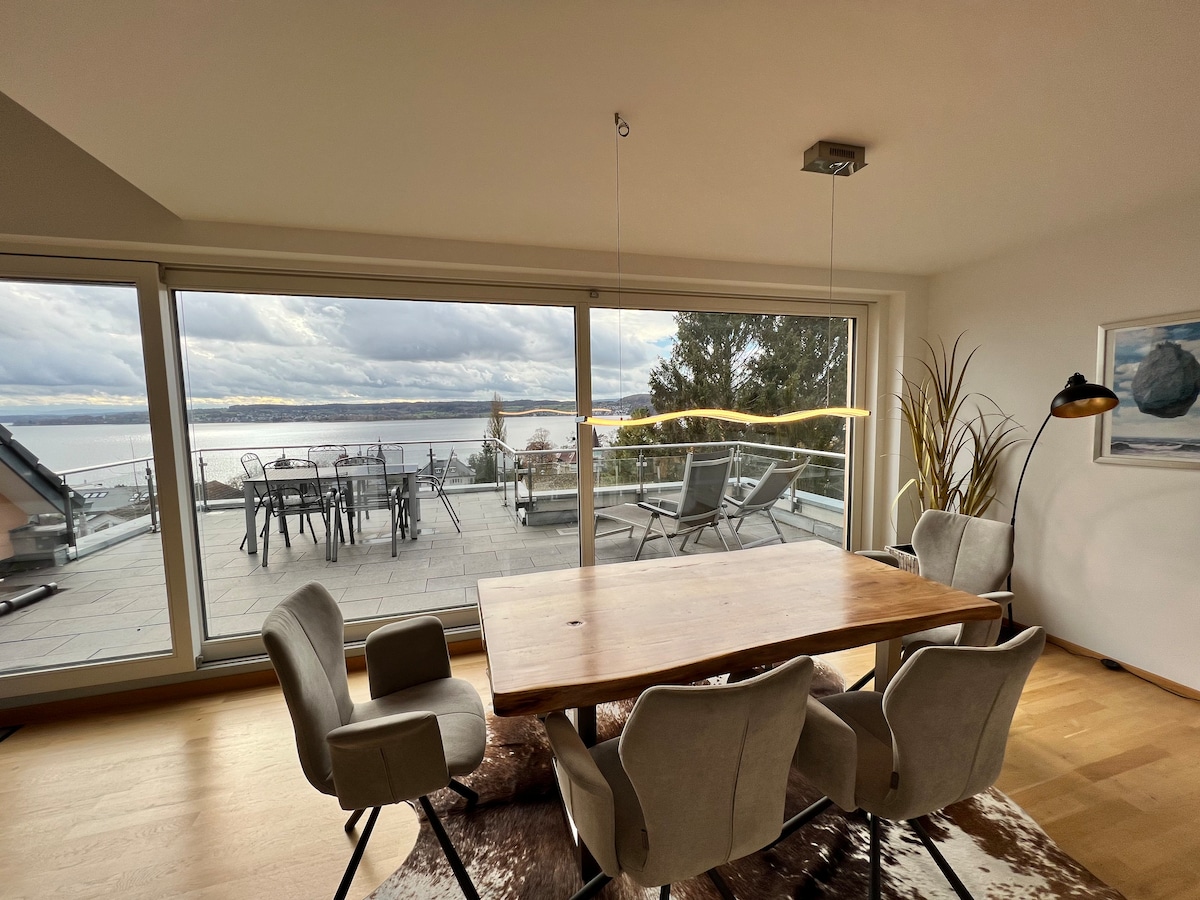 A dining area features a wooden table surrounded by comfortable chairs. Large glass doors open to a spacious terrace, offering a clear view of the lake. Natural light fills the room, complemented by contemporary decor and greenery in the corner.