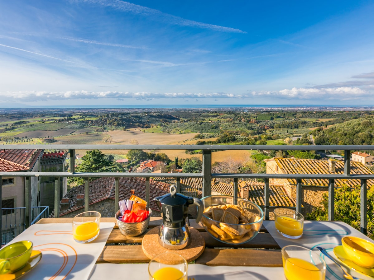 A table set for breakfast is displayed on a balcony, featuring a coffee pot, slices of bread, and fresh fruit. Glasses filled with orange juice complement the setting. In the background, a panoramic view of the countryside and sea is visible under a clear blue sky.