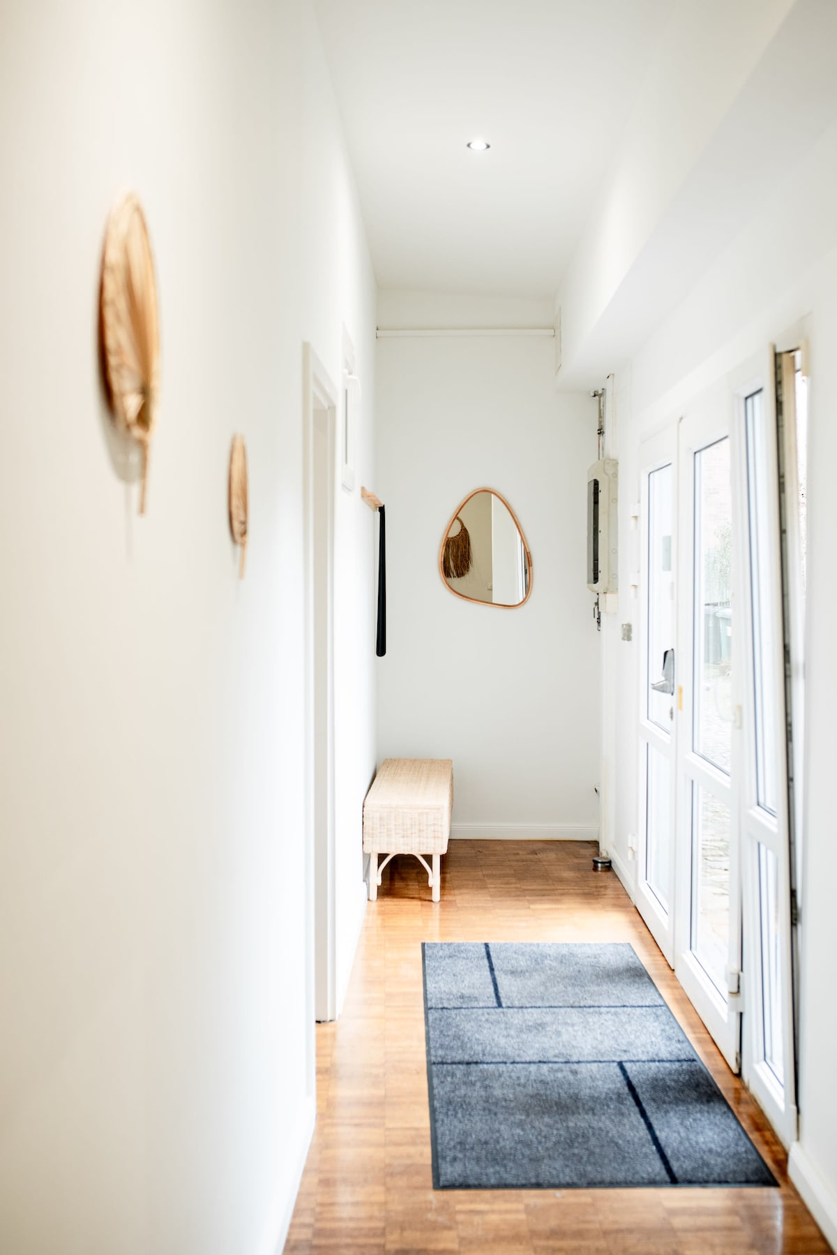 A clean and modern hallway features light-colored walls and polished wood flooring. A mirror is mounted on one side, reflecting the welcoming space. A simple bench is positioned against the wall, while a textured rug lies underneath, adding visual interest to the area.