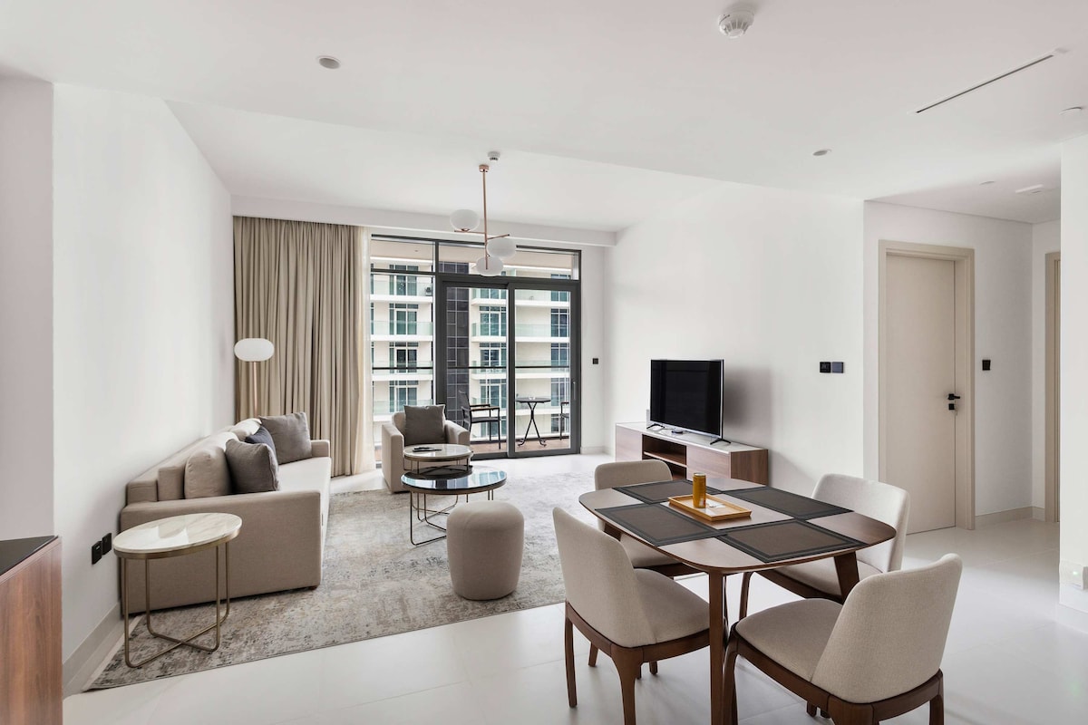 The open-plan living area showcases a modern design, featuring a comfortable sofa arrangement and a dining table with four chairs. A large window allows natural light to fill the space, offering access to a private balcony. A flat-screen TV is positioned across from the seating area.