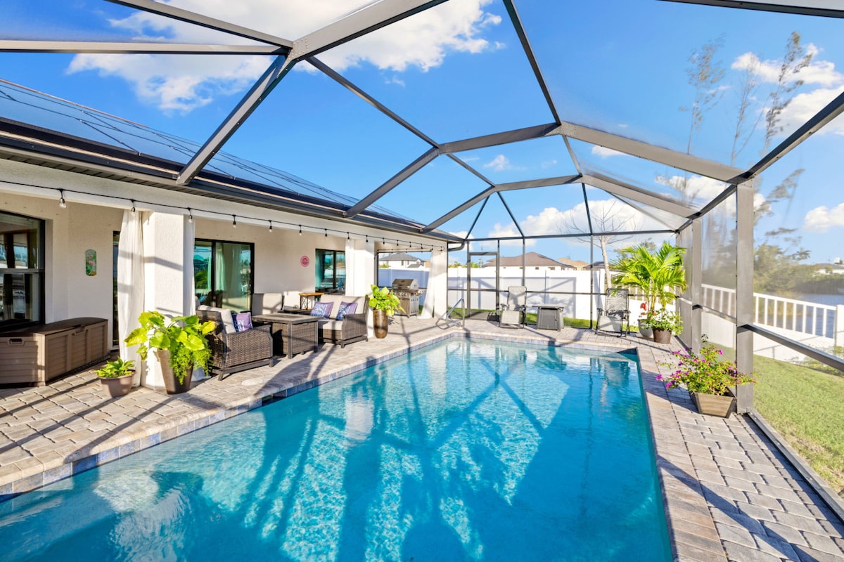 An outdoor area is showcased, featuring a heated pool surrounded by a spacious patio. Comfortable seating arrangements in dark wicker are present, complemented by vibrant potted plants. A clear view of the water is offered through the screened enclosure, with a bright blue sky overhead.