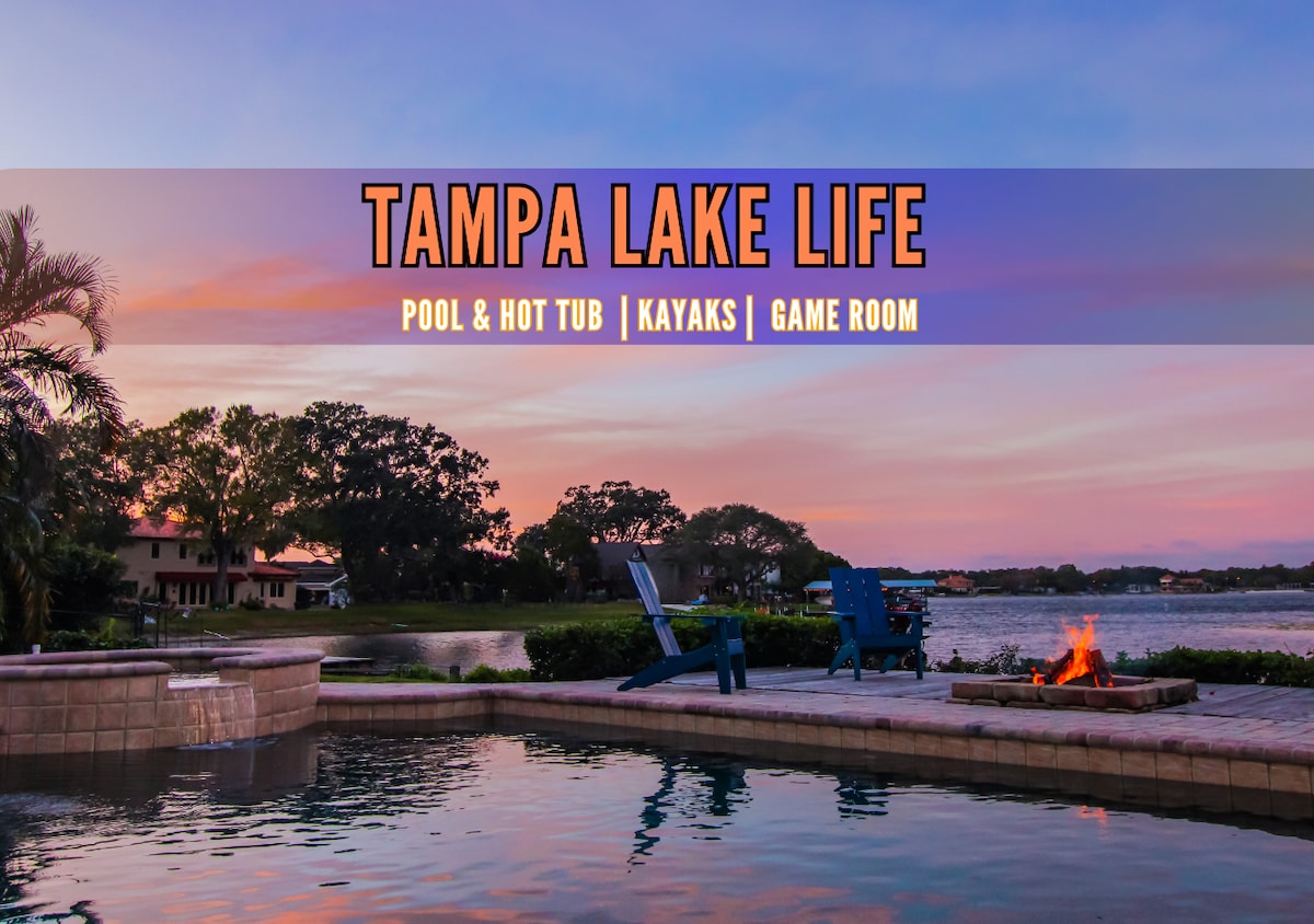 A serene waterfront view is presented, highlighting a private pool and hot tub beside a spacious patio. Adirondack chairs are positioned for relaxation, overlooking the lake. A fire pit adds warmth to the scene, framed by palm trees and a colorful sunset sky.