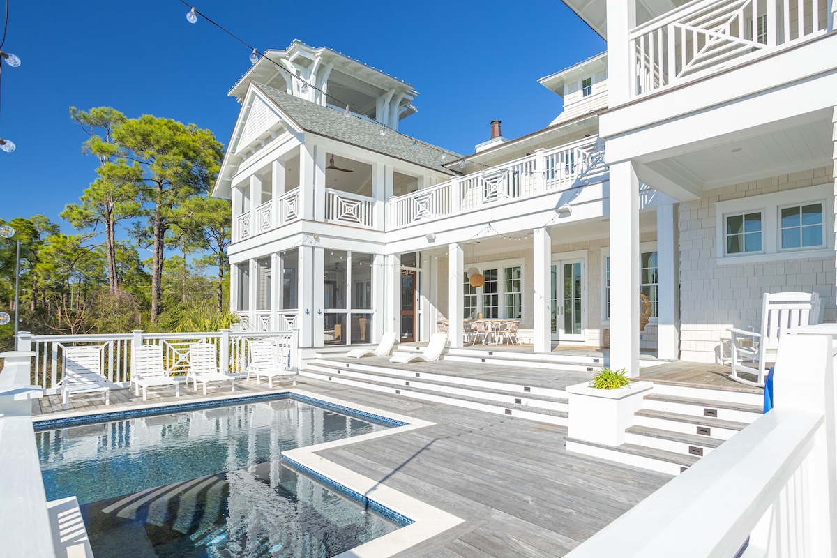 The exterior of a modern coastal home features a private heated pool surrounded by a spacious deck. White railings and outdoor seating enhance the area. Large glass doors connect interior spaces to the outdoor environment, set against a backdrop of lush greenery and clear blue skies.