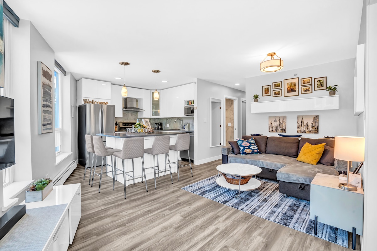 A modern living space is showcased, featuring a spacious sectional sofa with colorful cushions. A sleek kitchen with a large island and bar seating is visible in the background, illuminated by pendant lights. A light-colored area rug adds warmth to the room, complemented by framed artwork on the walls.