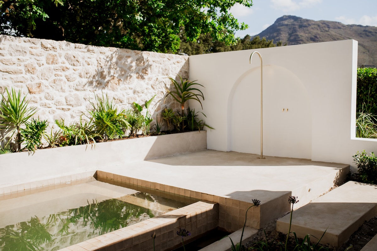 The outdoor area features a serene private pool, encompassed by smooth stone walls and lush greenery. A minimalist shower is set against a white arch, providing an inviting space for relaxation amid natural surroundings and distant mountain views.