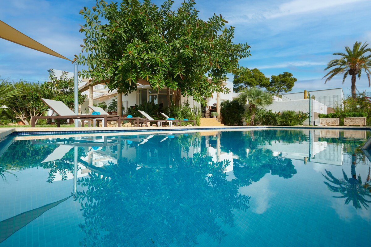 A spacious outdoor pool reflects the surrounding greenery and clear blue sky. Several lounge chairs are arranged around the pool, with the villa partially visible in the background, framed by lush trees and palm plants.