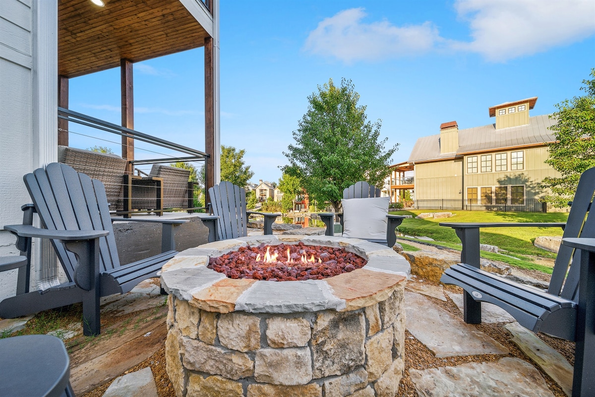 A stone firepit is surrounded by several dark wood chairs, inviting relaxation. Lush green landscaping and trees frame the background, with a picturesque view of the nearby cottage. The area is enhanced by a sense of outdoor comfort and communal gathering.