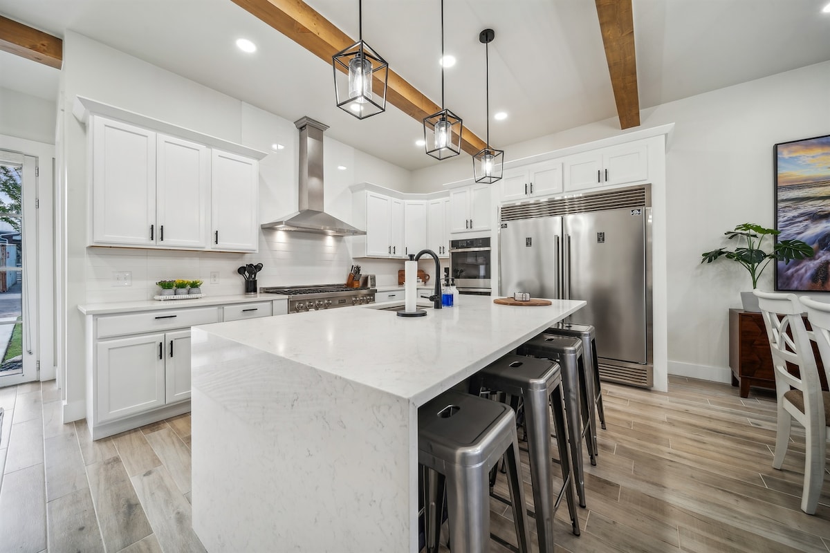 A modern kitchen features a spacious island with seating, complemented by sleek metal stools. Bright cabinetry and a stainless steel refrigerator are visible, along with a large, vented hood above the range. Warm wood beams extend across the ceiling, enhancing the inviting atmosphere.
