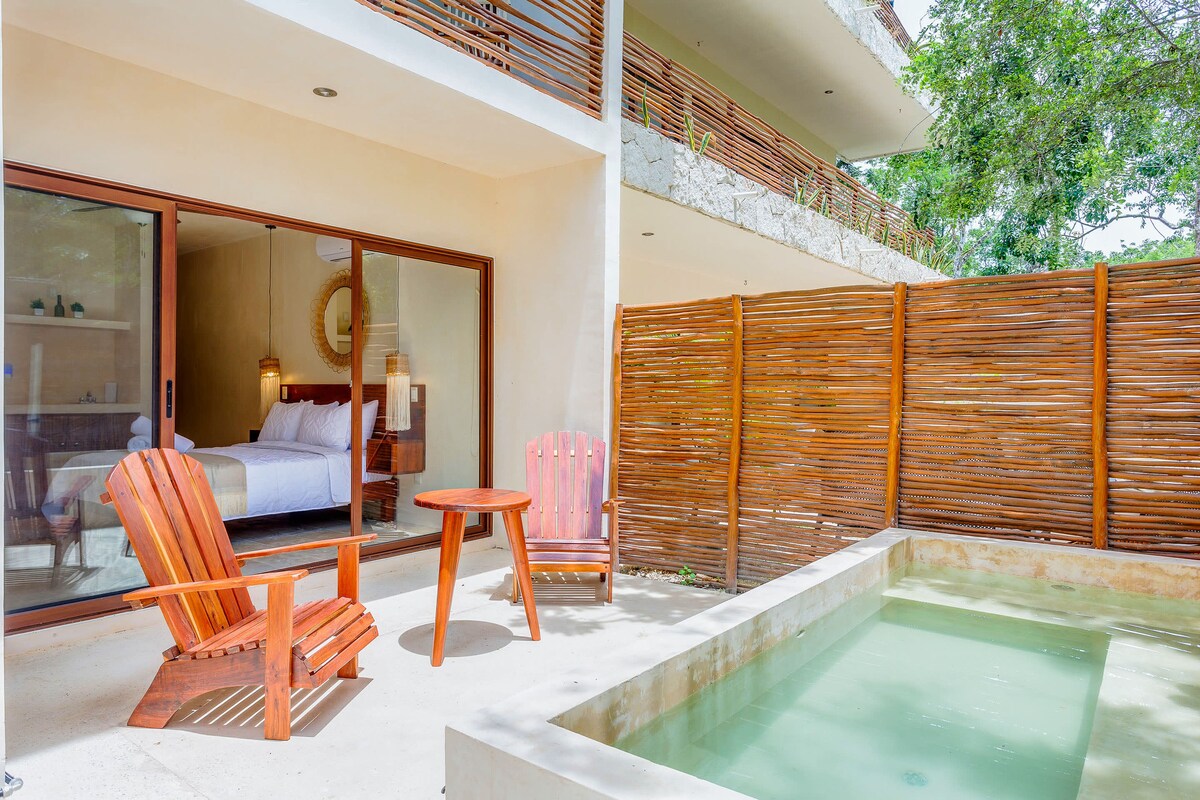 A private terrace features two wooden chairs and a small table, inviting relaxation. Glass doors provide a view into the bedroom with a spacious bed. A small plunge pool adds to the serene outdoor ambiance, surrounded by natural wood fencing.