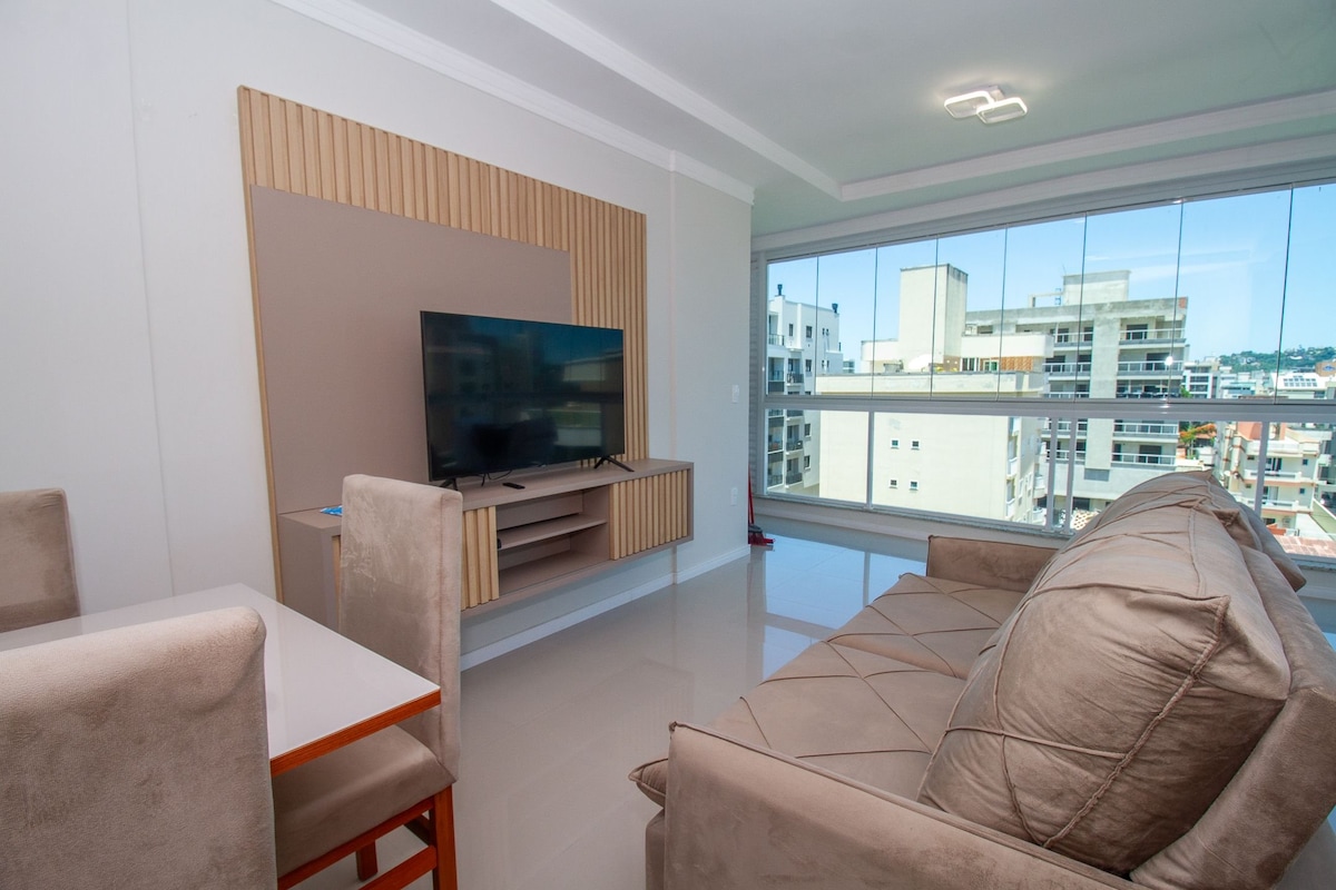 A modern living area features a comfortable sofa facing a mounted flat-screen television. A dining table for six is positioned nearby, while large windows allow natural light to fill the space, offering a view of the surrounding neighborhood.
