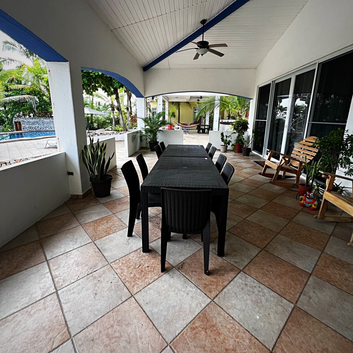 A spacious covered patio features a large rectangular dining table surrounded by six chairs. The tiled floor is complemented by green plants scattered throughout, adding a touch of nature. Views of the pool and garden area can be seen through large glass doors.