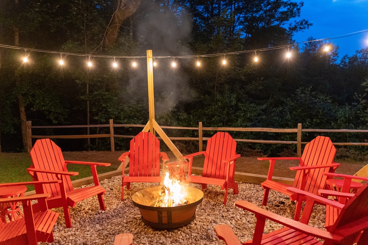 A cozy fire pit area is surrounded by six red Adirondack chairs positioned on gravel. Soft cafe lights hang above, casting a warm glow, while smoke rises gently from the crackling fire, set within a circular metal pit. Lush greenery is visible in the background.