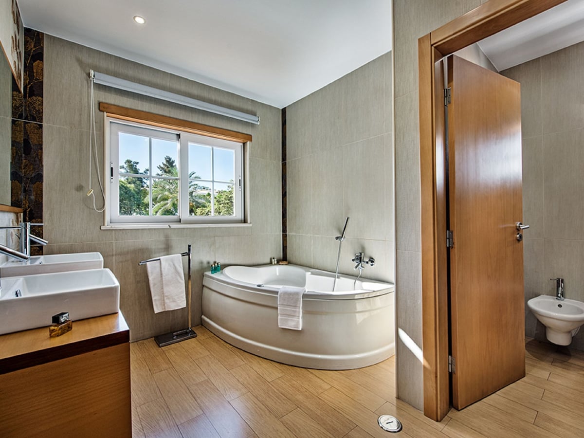 A well-lit bathroom features a curved bathtub, along with two modern sinks positioned on a wooden vanity. A window allows natural light to flow in, offering views of greenery outside. A bidet is also visible, and warm-toned tiles cover the floor.