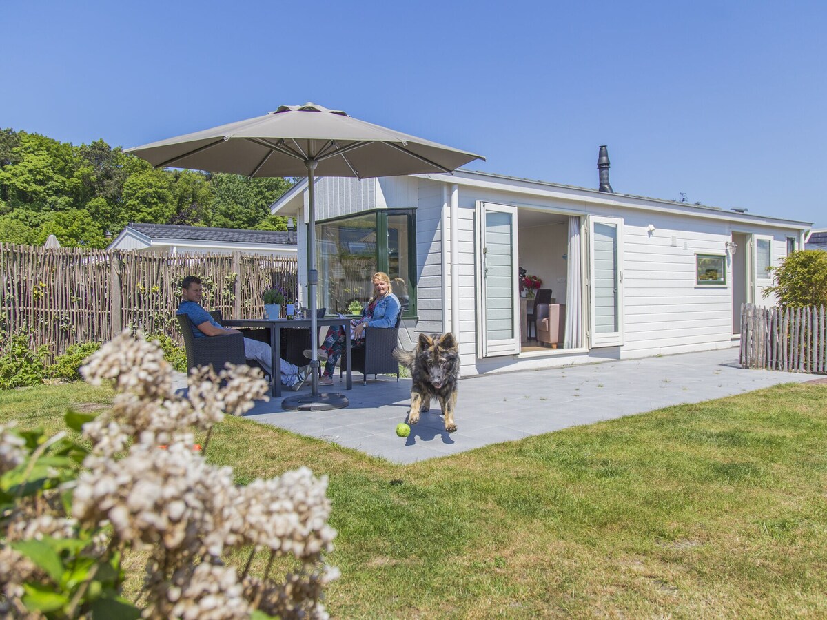 A charming terrace area is presented outside the chalet, complete with an umbrella over a table and chairs. A grassy space extends towards a fence, while a dog is seen playing with a tennis ball nearby. The open doors of the chalet invite natural light into the interior.