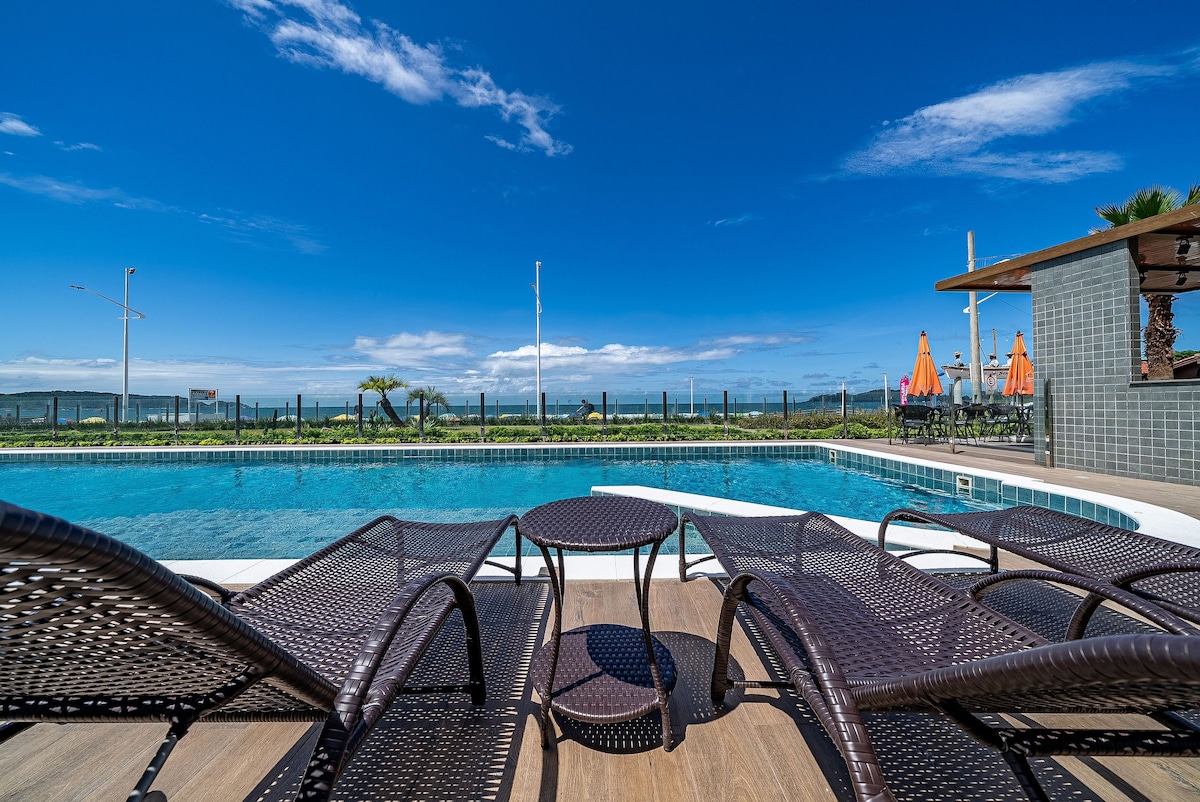 A tranquil swimming pool is surrounded by lounge chairs and a small table, offering views of the expansive sky and greenery beyond. Clear blue waters reflect the sunlight, inviting relaxation in this open outdoor space.