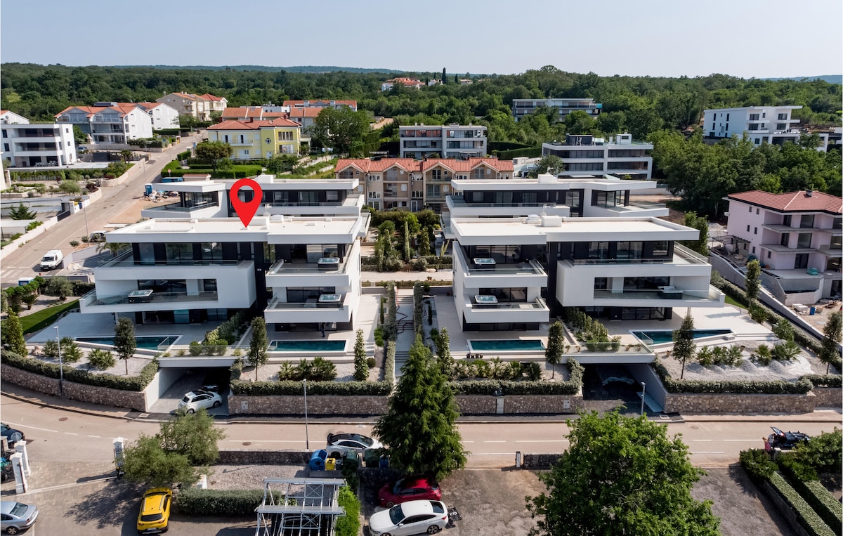 Aerial view of a modern residential complex, showcasing multiple contemporary buildings with flat roofs and balconies. Lush greenery surrounds the properties, and several swimming pools can be seen. The layout provides easy access to outdoor spaces, with streets lined by trees and parked cars.