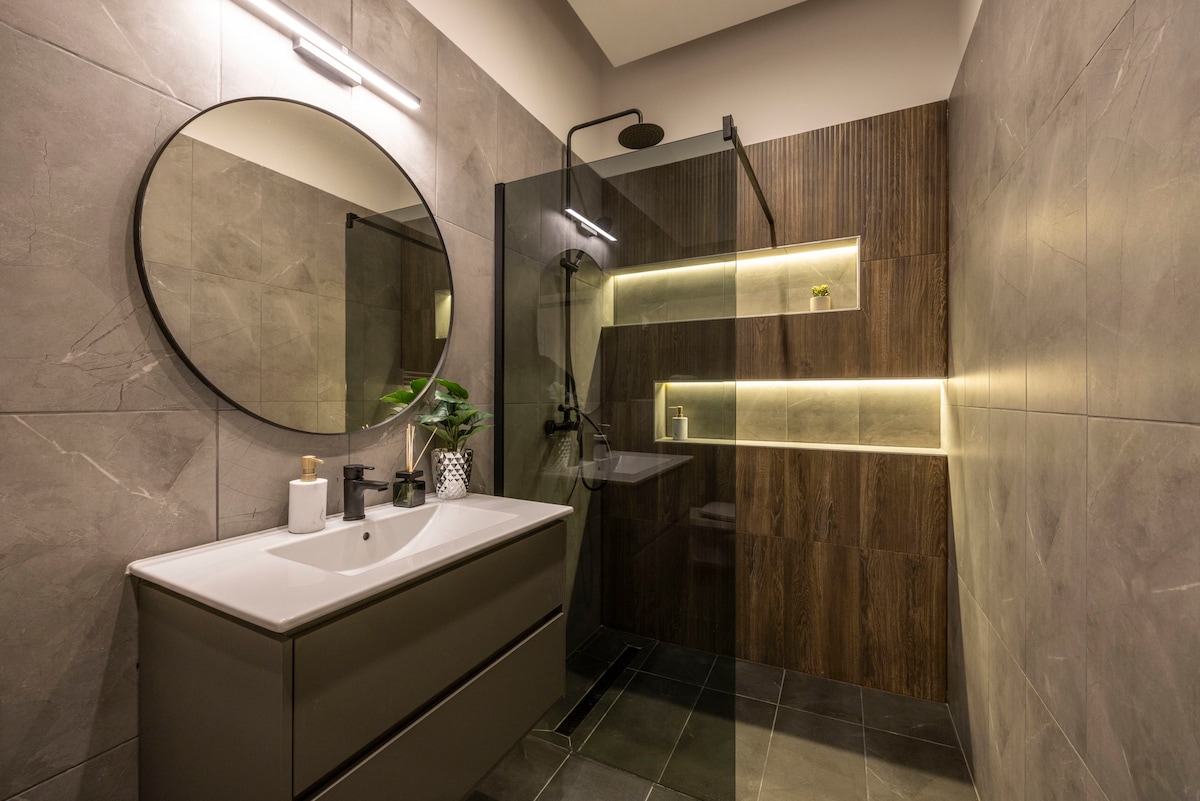 A modern bathroom features a round mirror above a sleek vanity with a light-colored countertop. The shower area is enclosed with glass, showcasing a rainfall shower head. Soft lighting highlights the textured wall, which combines stone and wood elements, creating a contemporary atmosphere.