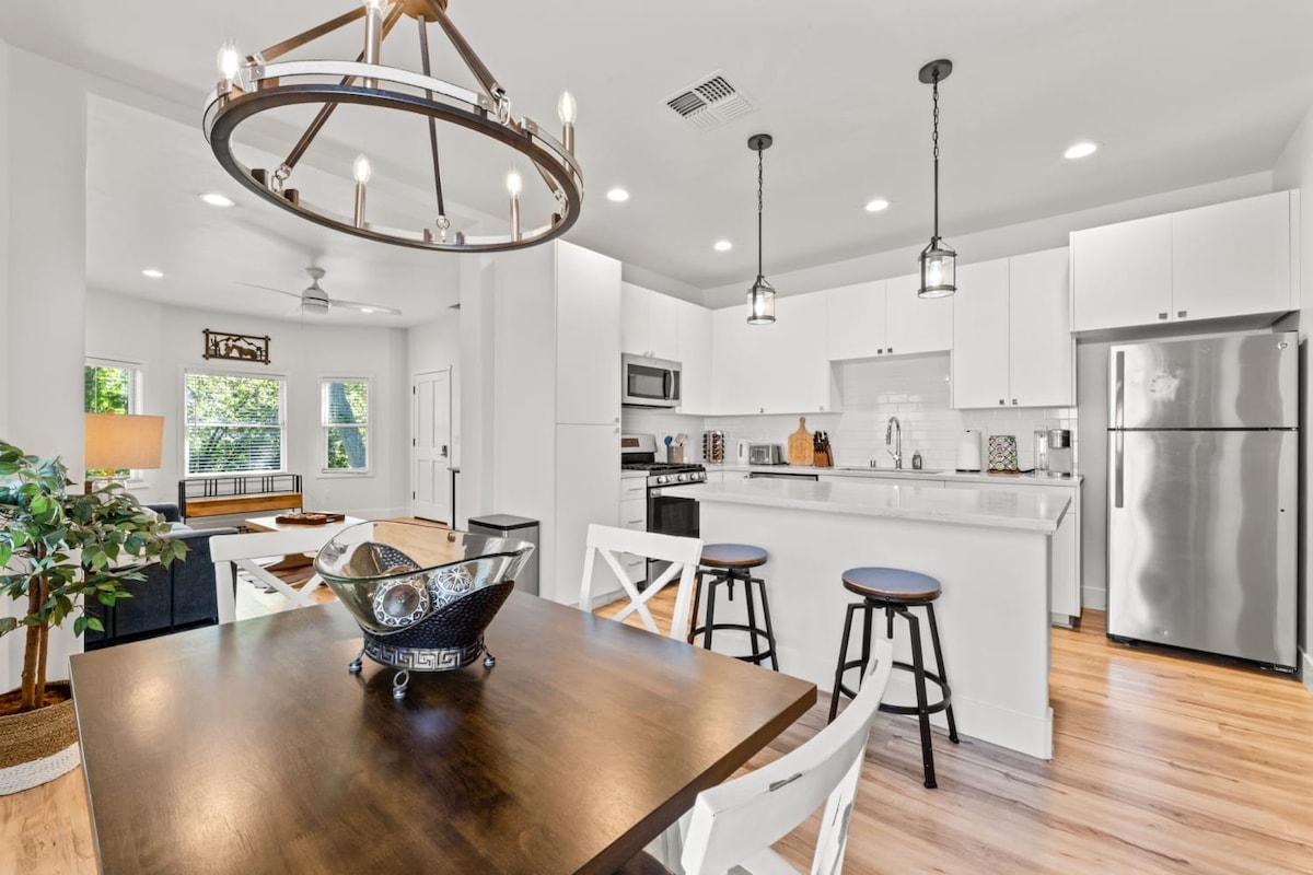 An open-concept kitchen and dining area features modern appliances, including a stainless steel refrigerator and oven. A large dining table is accompanied by white chairs, while a stylish chandelier provides overhead lighting. Natural light fills the space through multiple windows, enhancing its bright ambiance.