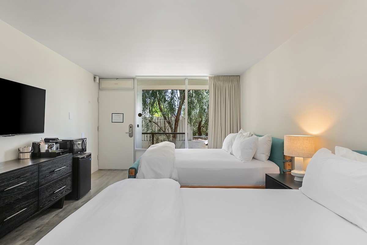 A bright hotel room featuring two spacious beds dressed in crisp white linens. A flat-screen TV is mounted on the wall, and a dark wooden dresser holds refreshments. Large sliding glass doors lead to an outdoor area, with natural light illuminating the space.