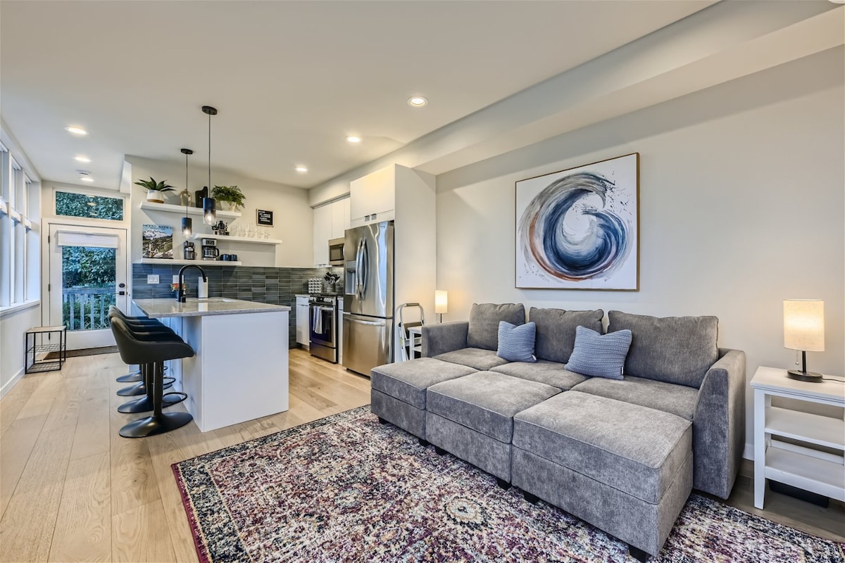 The living area is furnished with a spacious gray couch, accompanied by blue accent pillows. High-top seating at a breakfast bar enhances the kitchen's modern design, which features stainless steel appliances and light cabinetry. Natural light filters through large windows, illuminating the room.