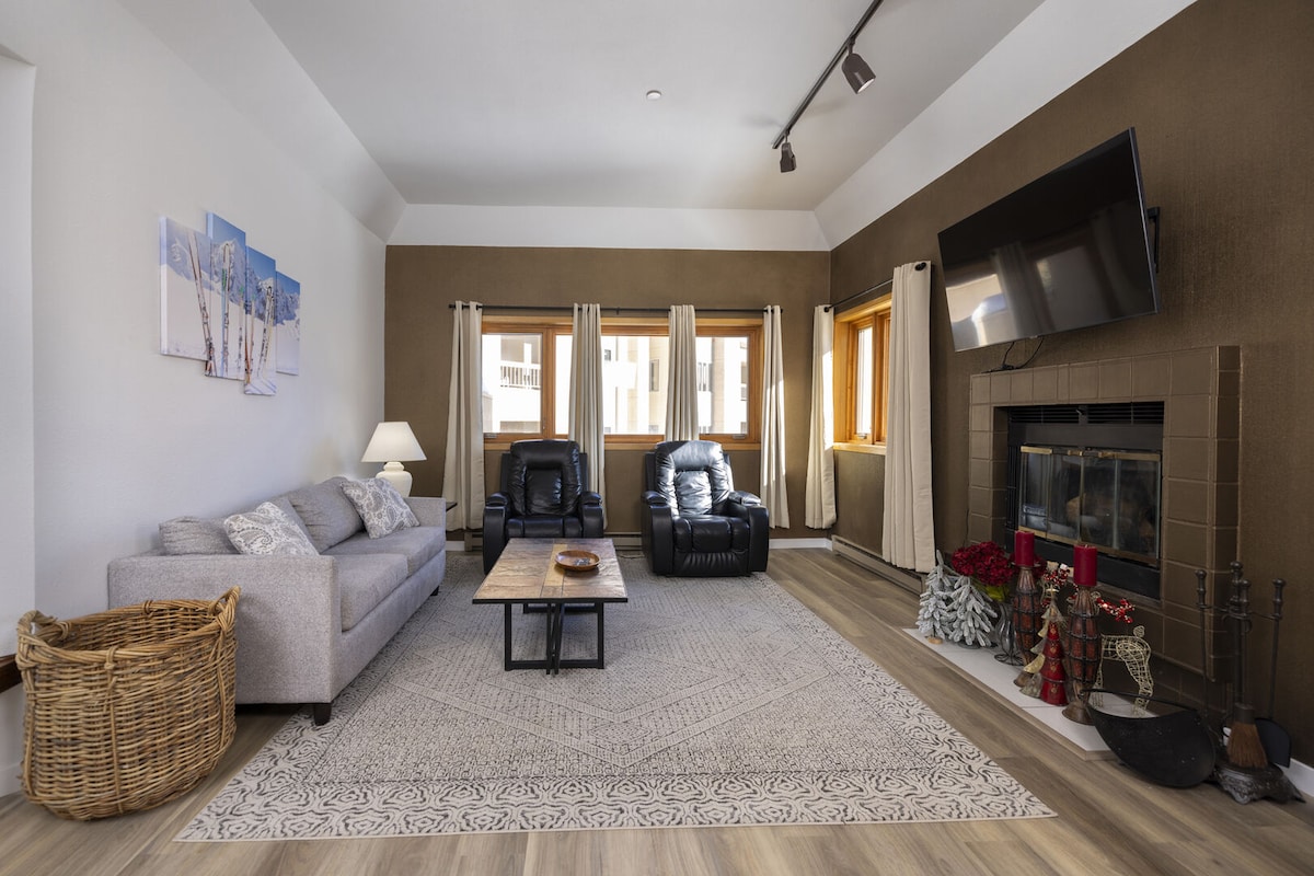 A spacious living area is highlighted by a light-colored sofa and two black armchairs. A coffee table sits in the center, with a fireplace adorned with seasonal decorations nearby. Natural light filters through the large windows, framed by soft drapes.