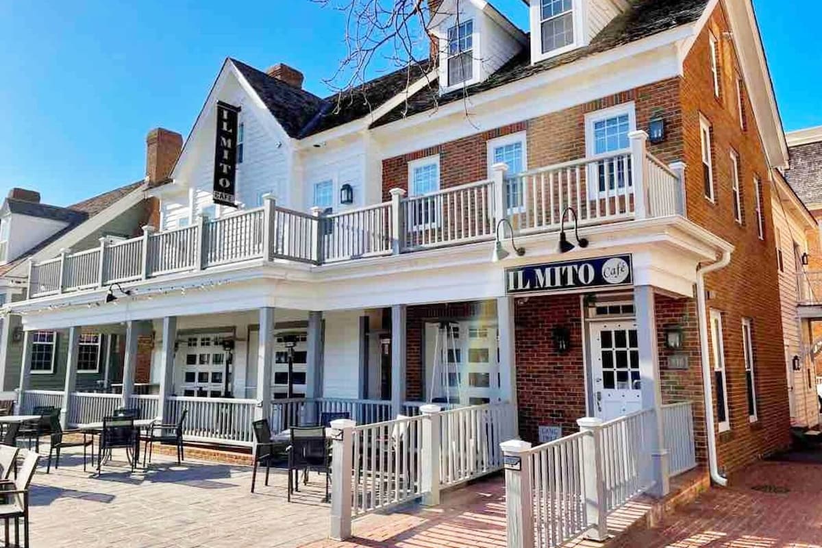 A charming two-story building features classic colonial architecture, with white and brick facades. A spacious outdoor seating area is visible, adorned with black metal tables and chairs. Balconies with white railings extend along the front, inviting guests to relax and enjoy the surroundings.