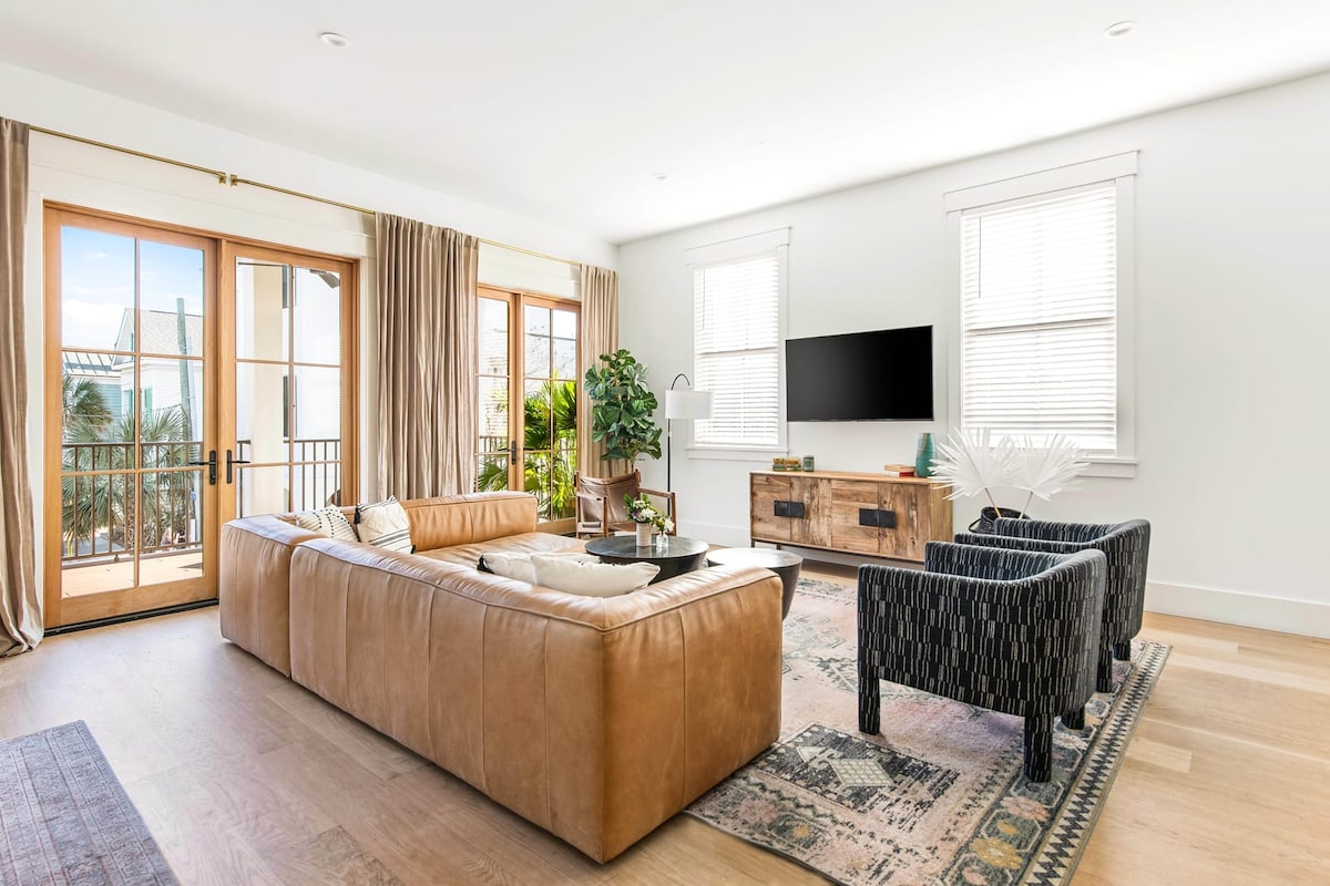 The living area is characterized by a large leather sectional sofa and two patterned armchairs. A sleek media console stands beneath a mounted TV. Sunlight streams through glass doors leading to a balcony, with lush greenery adding a touch of nature to the space.
