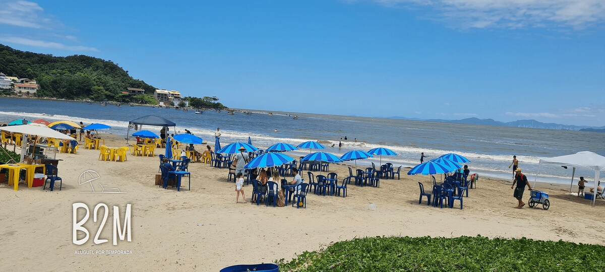 An inviting beach scene features a sandy shore dotted with umbrellas and chairs in blue and yellow. The gentle waves of the ocean stretch into the distance, framed by lush green hills under a clear blue sky. Beachgoers can be seen enjoying the sunny day.