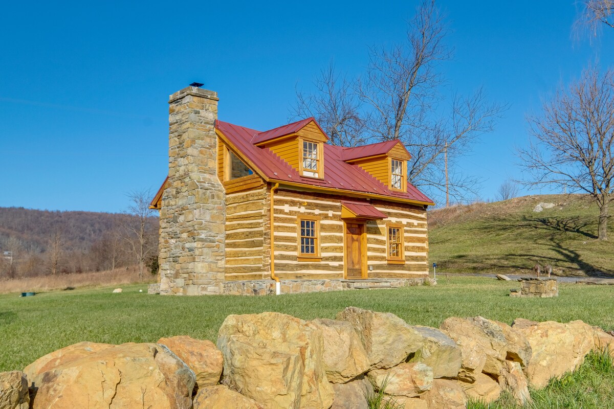 A rustic cabin is showcased with wooden log construction and a red metal roof. A stone chimney rises prominently beside the entrance. The cabin is set against a backdrop of rolling green hills and a clear blue sky, creating a serene countryside ambiance.