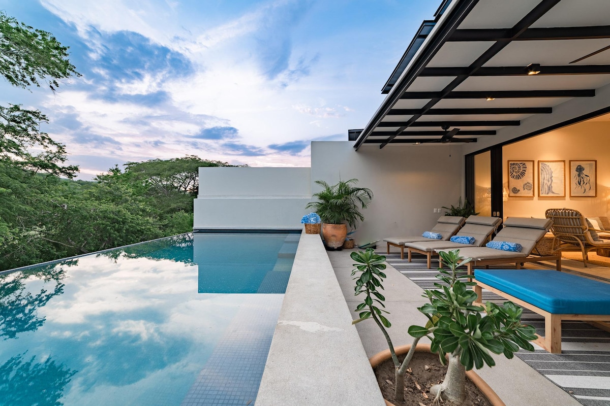 An expansive pool terrace is visible, showcasing a sleek infinity pool that blends with the tropical landscape. Comfortable loungers are arranged alongside, while the backdrop features large windows revealing the interior living space. Lush greenery surrounds the area, complementing the tranquil setting.