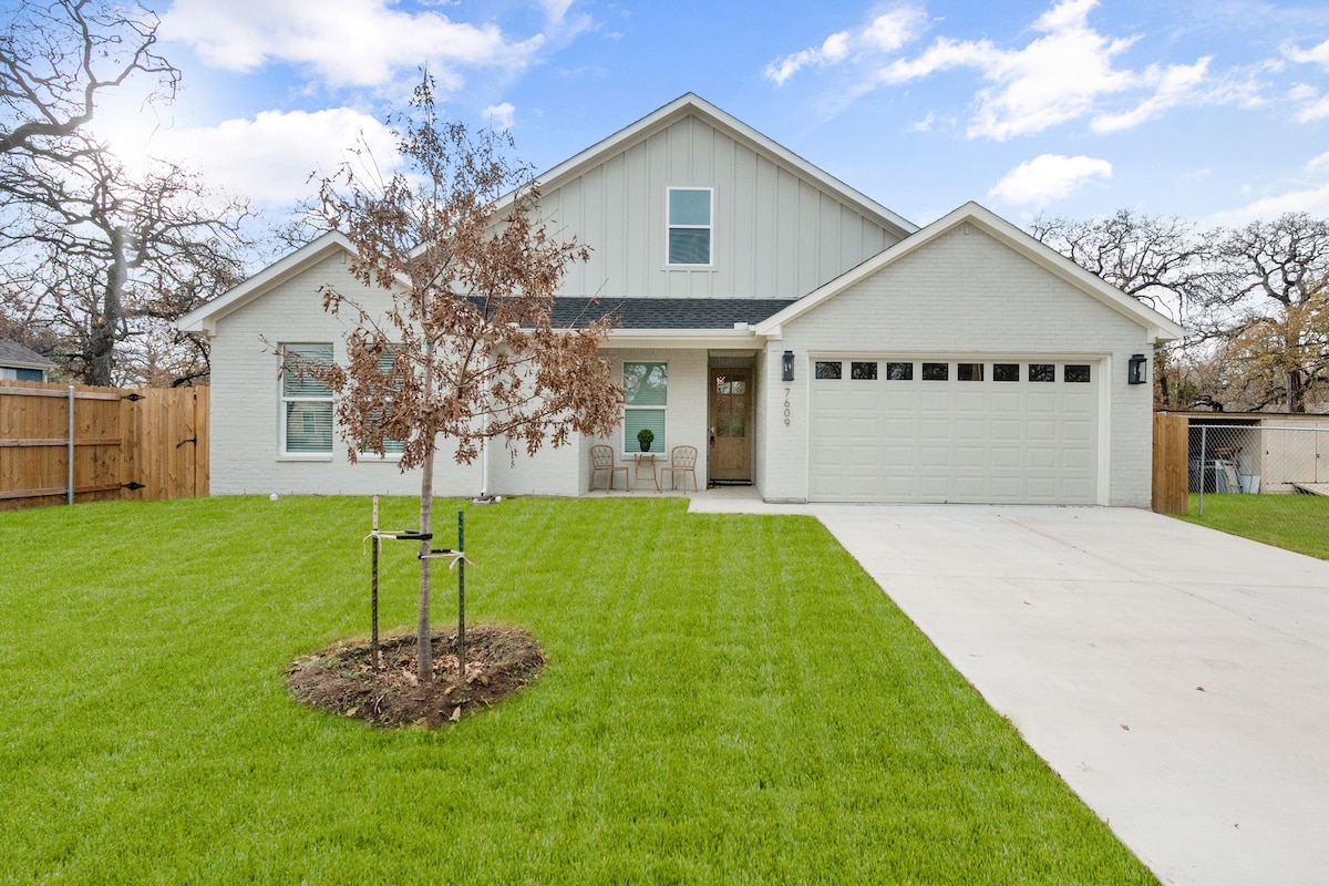 A modern single-story home is centered on a well-manicured lawn, featuring a symmetrical facade with light-colored brick. Two chairs are positioned near the entryway, while a small, newly planted tree is visible in the foreground. The driveway leads to a spacious garage.