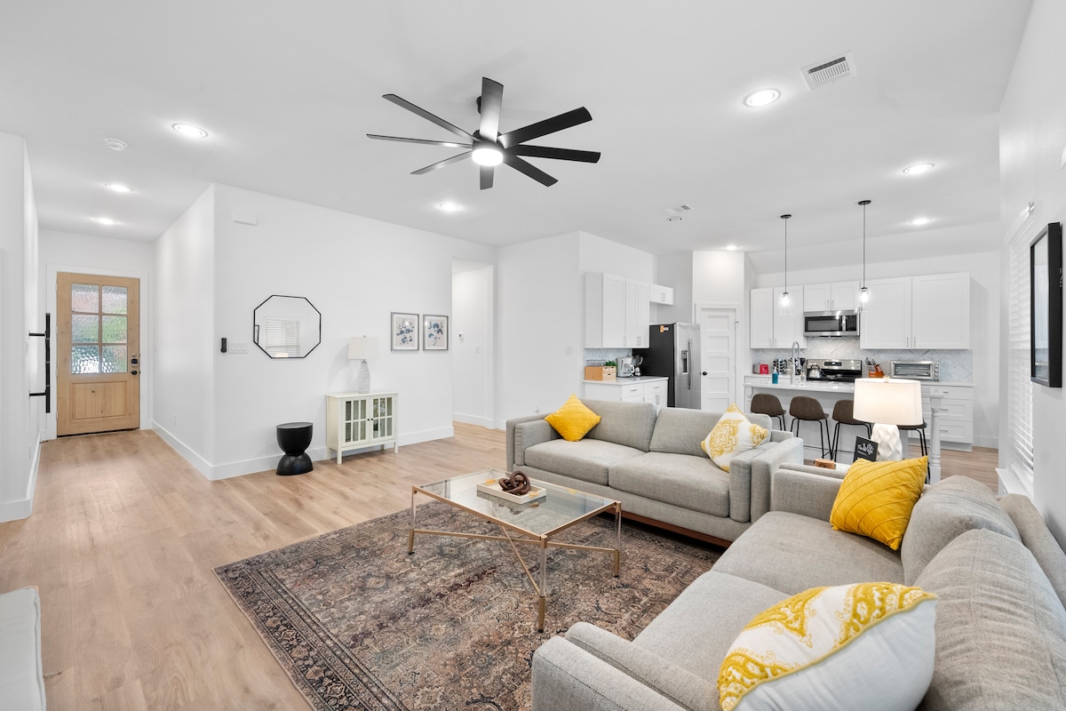 A spacious common area features modern furnishings, including a light gray sectional sofa adorned with a yellow accent pillow. A glass coffee table is centered on a decorative rug. The open layout allows natural light to flow in from the adjacent kitchen and entryway.