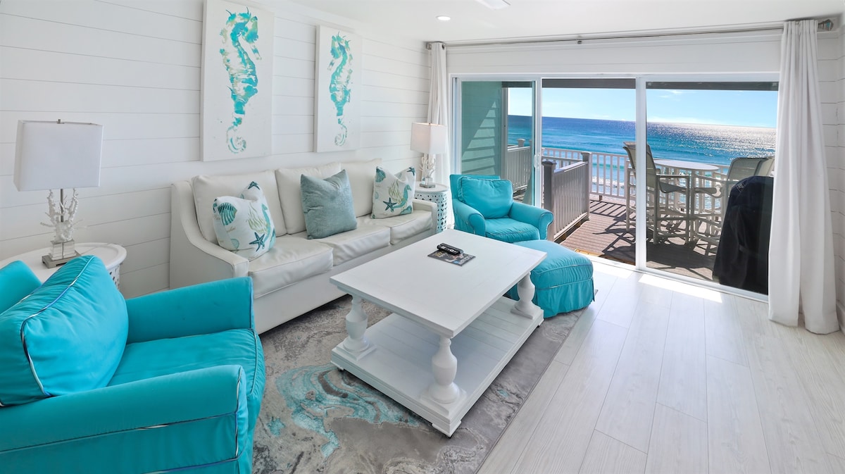 A bright living area features a white sofa and accent chairs in turquoise. A coffee table sits in the center, surrounded by a patterned rug. Large sliding doors reveal a stunning view of the beach, leading to an outdoor seating area.