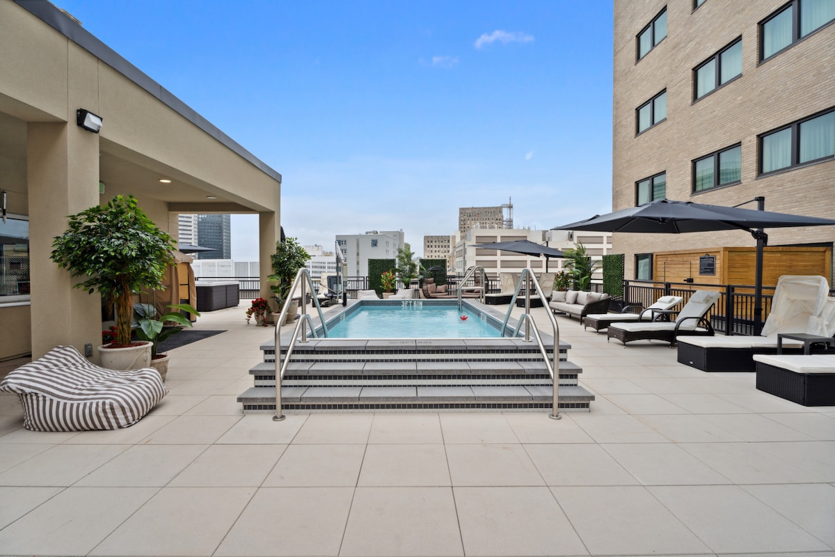 The rooftop pool area features a large swimming pool surrounded by comfortable lounge chairs and umbrellas. Large planters with greenery provide a touch of nature. The area is paved with light-colored tiles, offering ample space for relaxation under a clear blue sky.