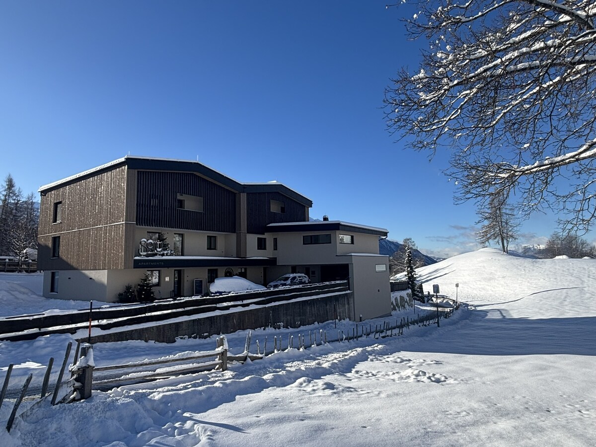The building features a modern design with a combination of dark wood, light-colored walls, and large windows. Surrounding landscapes are coated in fresh snow under a clear blue sky, providing a serene winter atmosphere. Snow-covered hills can be seen in the background.