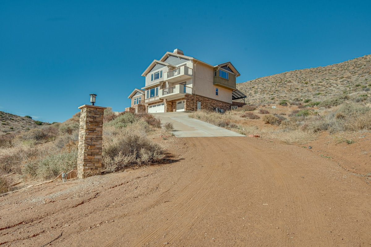 The multi-story home is positioned on a gentle slope, surrounded by natural landscaping. A stone pillar marks the driveway leading to the entrance, while the building boasts a mix of stucco and stone elements under a clear blue sky.