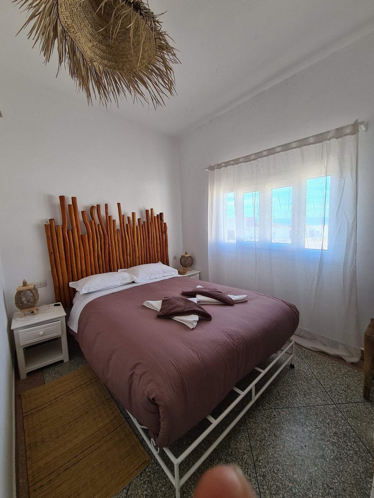 A cozy bedroom features a natural wood headboard and soft bedding in muted tones. Light pours through a large window, illuminating the room, while a bedside table and decorative elements add a touch of warmth and character.