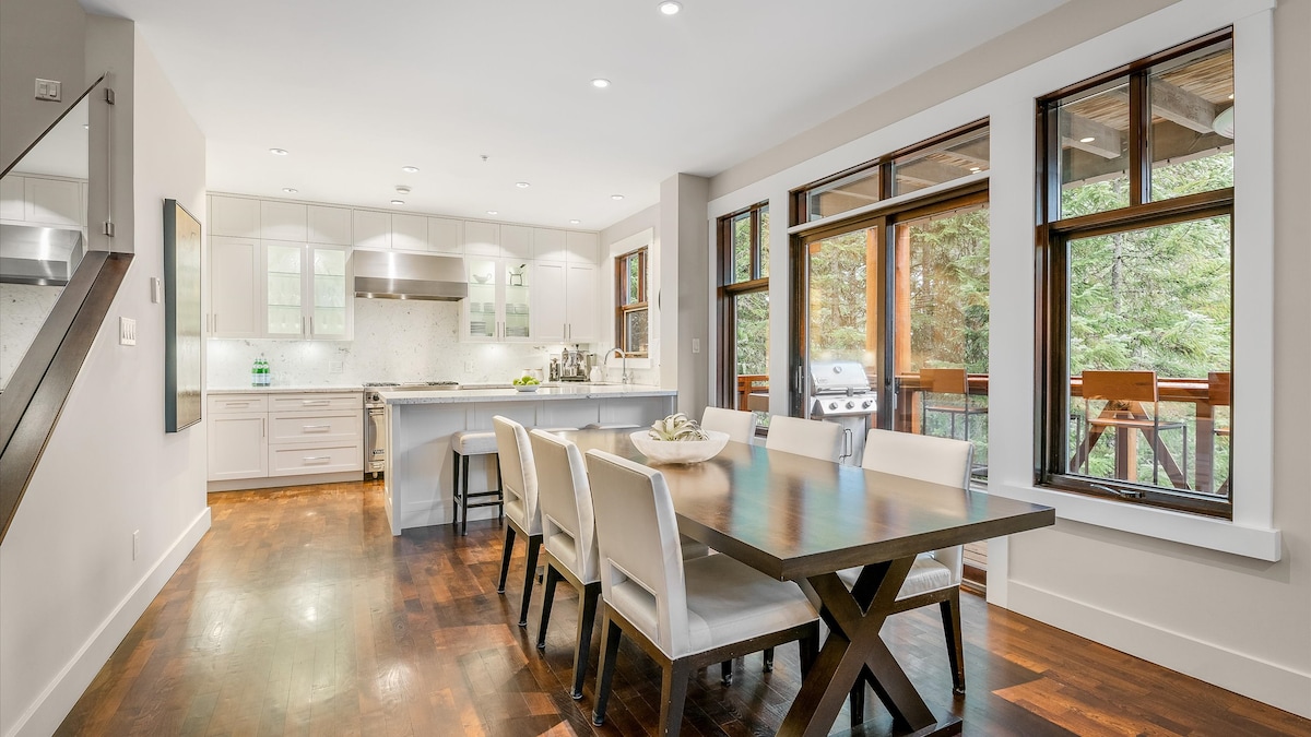 The spacious kitchen and dining area feature modern cabinetry and stainless steel appliances. Large windows allow natural light to fill the room, offering views of the surrounding forest. A large dining table comfortably seats six, complemented by elegant chairs and a decorative centerpiece.