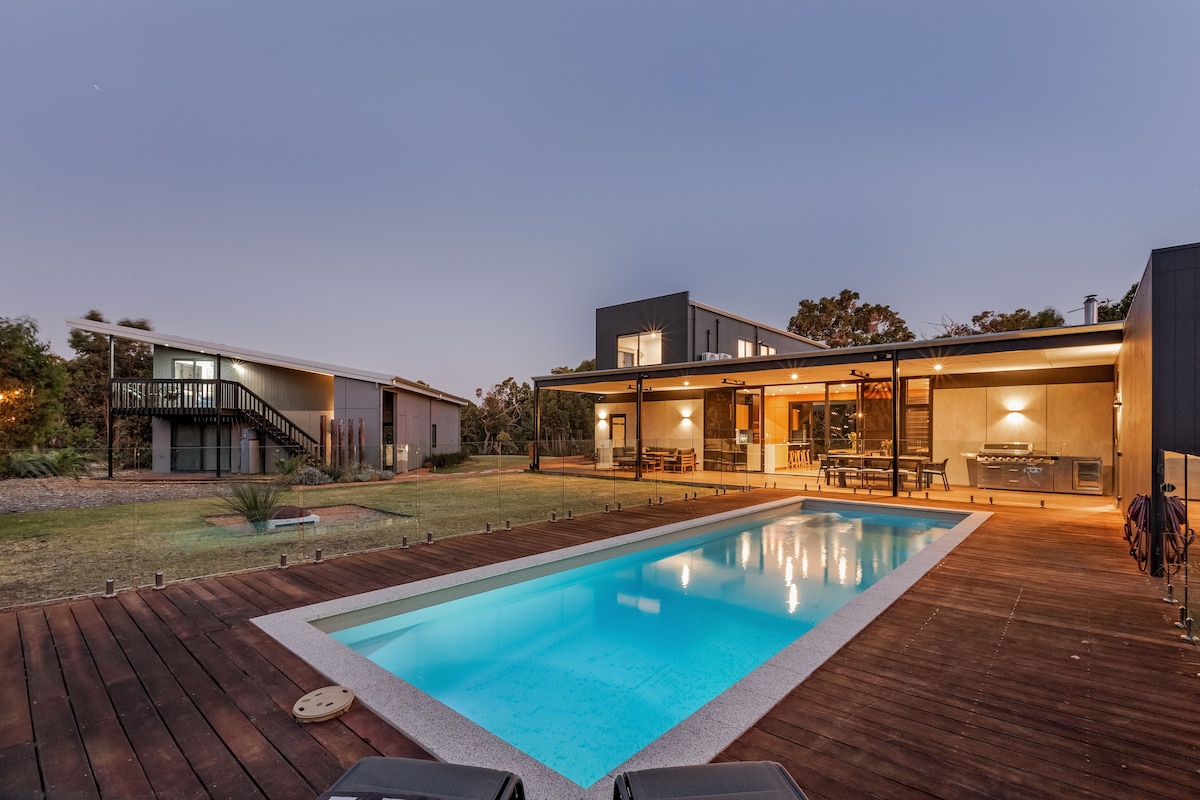 A spacious outdoor area features a shimmering pool surrounded by timber decking. The modern two-story building displays large glass windows and sliding doors, providing views of lush greenery and sleek outdoor furniture, ideal for relaxation and entertaining.