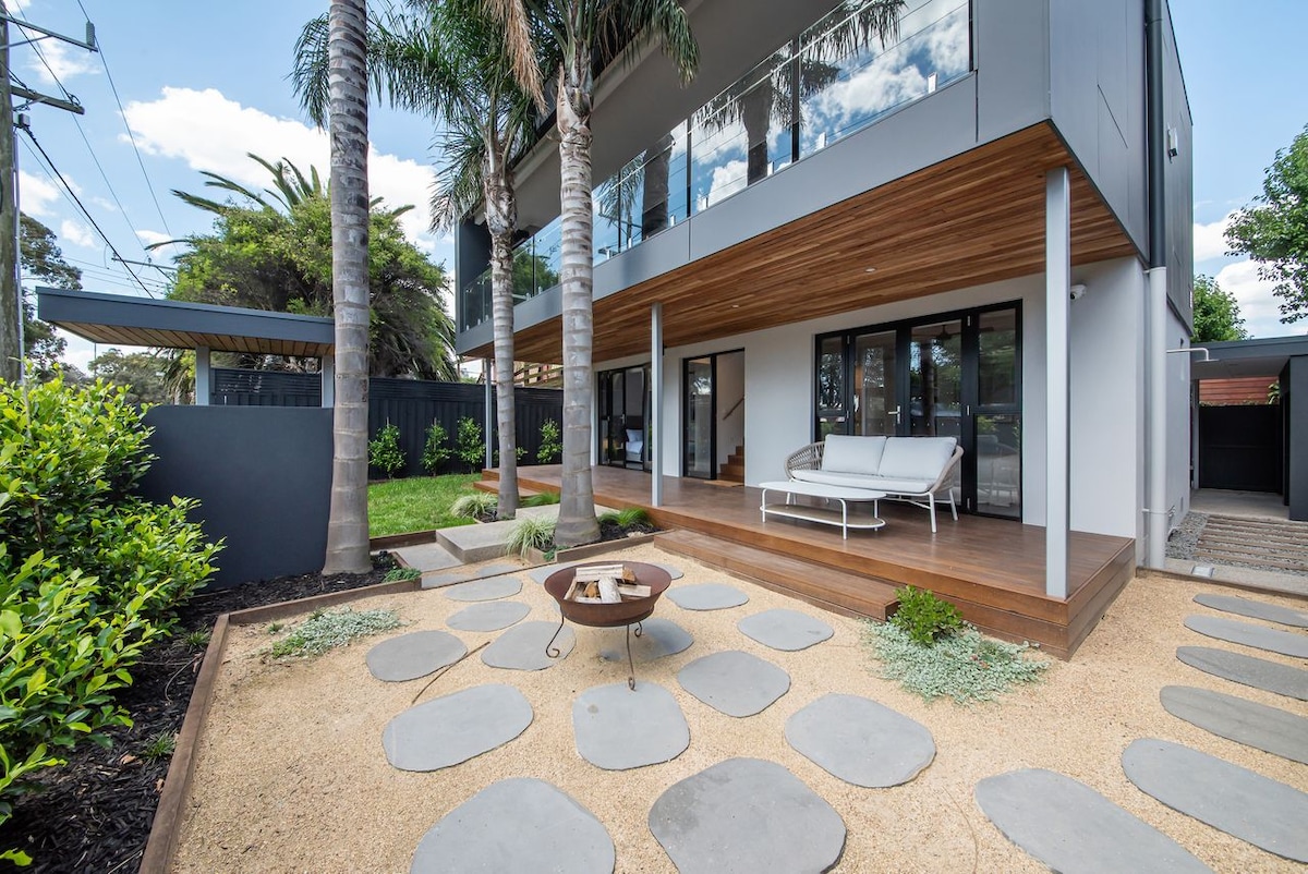 The outdoor area features a wooden deck with a comfortable seating arrangement. A circular fire pit sits on a path of large stone pavers surrounded by gravel and greenery. Palm trees provide shade, enhancing the inviting outdoor atmosphere.