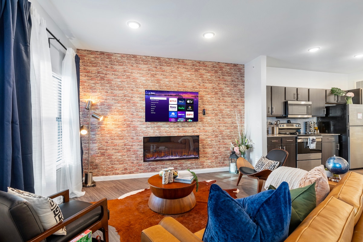 The living area features a stylish sofa arranged around a round coffee table, complemented by an accent rug. A smart TV is mounted on a brick accent wall, with soft lighting illuminating the room. A large window with sheer curtains allows natural light to brighten the space.