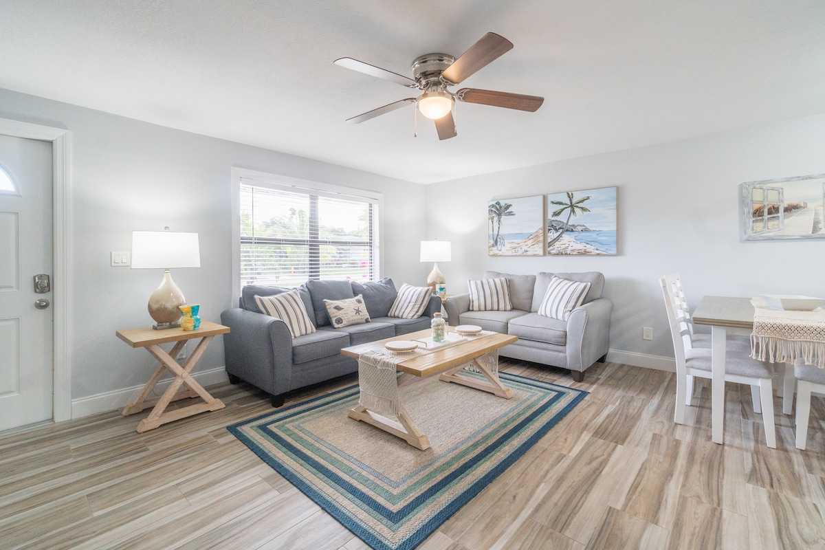 A well-lit living area features a gray sofa complemented by decorative pillows. A wooden coffee table is centered on a patterned area rug, surrounded by two side tables with lamps. Light-colored dining table and chairs are positioned nearby, enhancing a cohesive and welcoming atmosphere.
